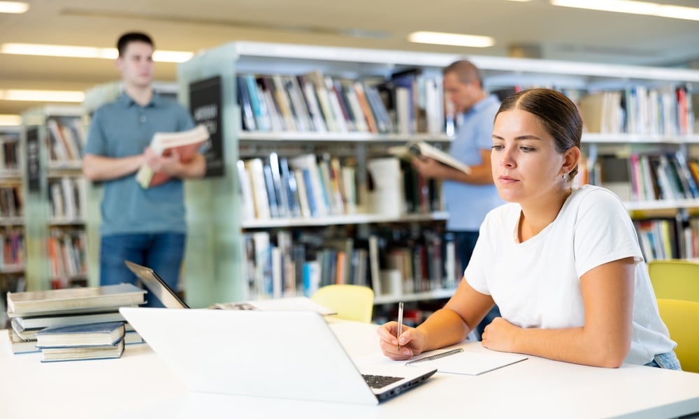 Étudiante concentrée prenant des notes devant son ordinateur dans une bibliothèque calme entourée de livres.