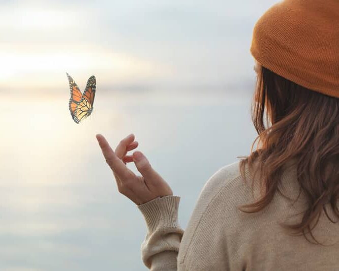 Papillon monarque volant près d'une femme au bord de l'eau Femme en bonnet orange admirant un papillon monarque volant au-dessus de sa main près de l'eau au coucher du soleil.