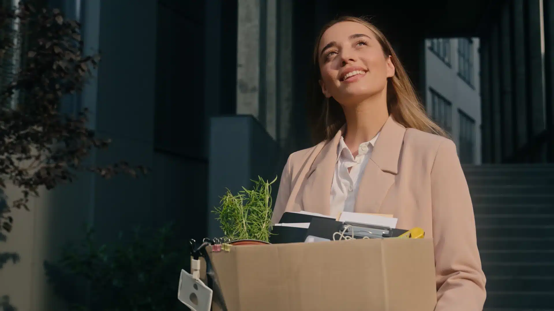 Jeune femme souriante portant un carton de bureau avec une plante, illustrant une démission ou un nouveau départ.
